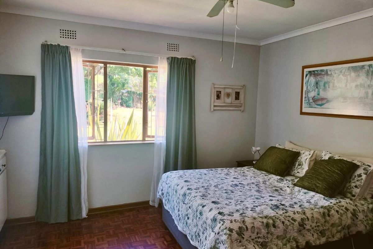 A well-lit bedroom featuring classic parquet flooring, a ceiling fan, and a window with security bars overlooking the garden.