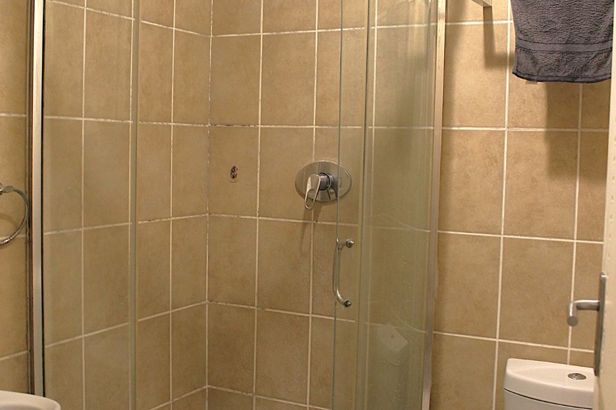 A compact bathroom featuring a curved glass shower enclosure, ceramic tiling, and a standard toilet, showing signs of wear and dated finishes.