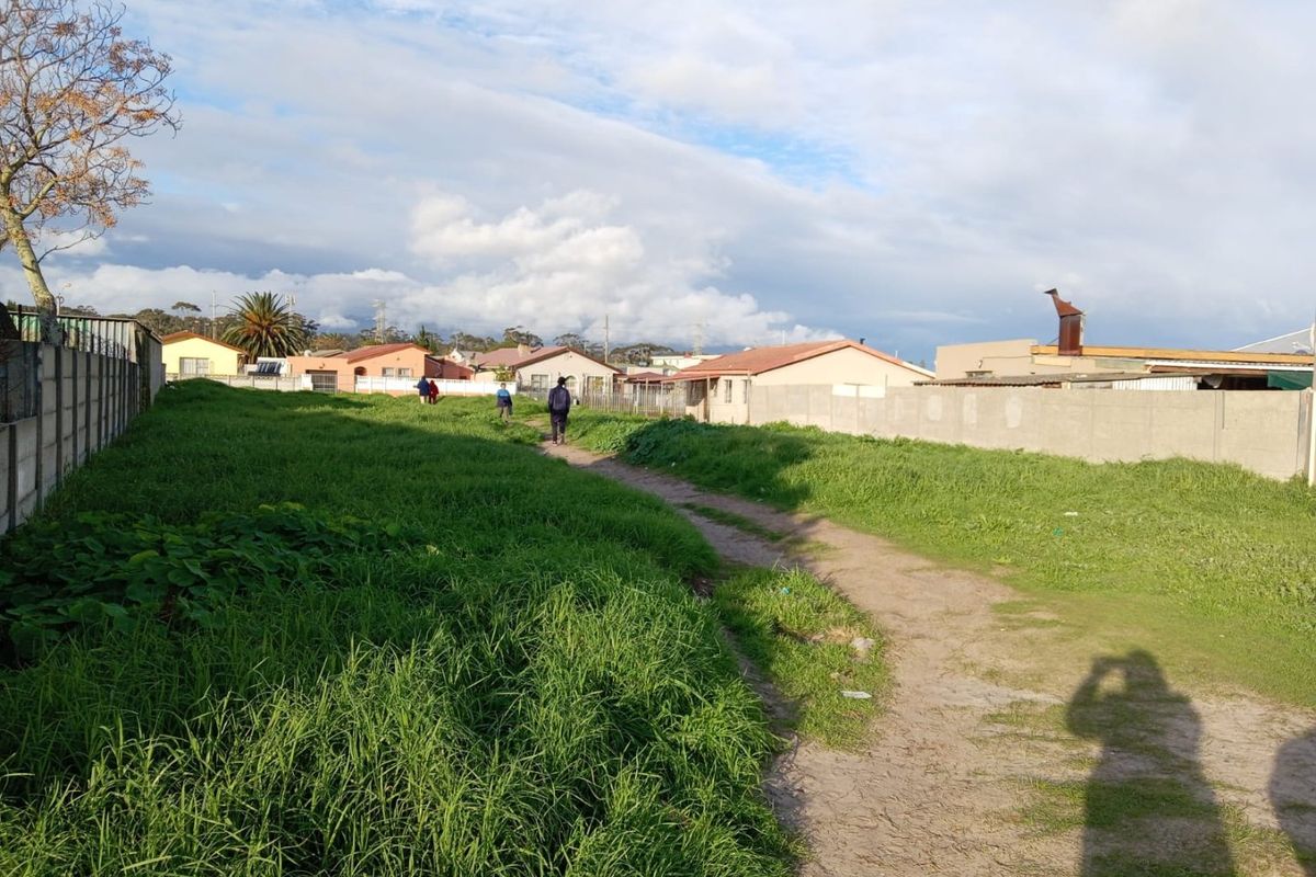 A view of a vacant residential plot featuring a dirt path and perimeter boundary walls, surrounded by neighboring residential properties.