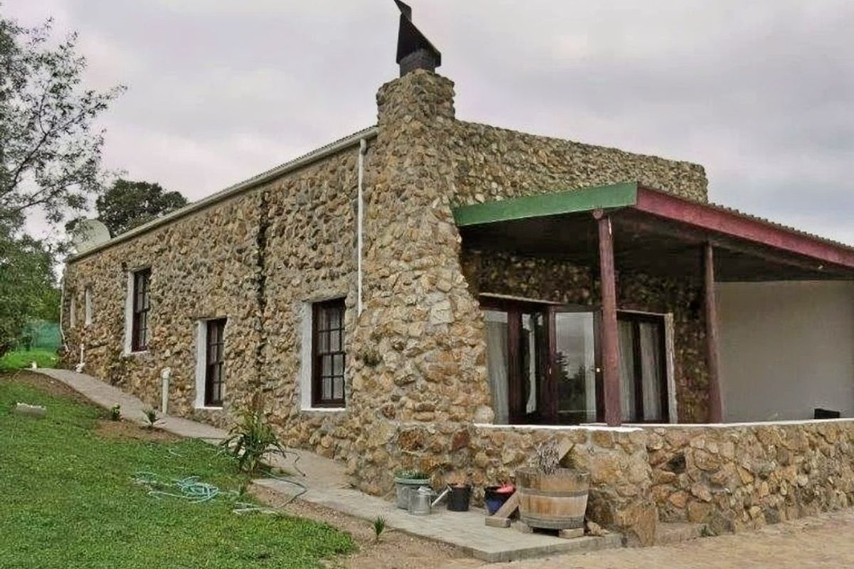 A rustic stone-clad house exterior featuring a covered patio area, paved walkway, and a prominent chimney stack set within a grassy garden.