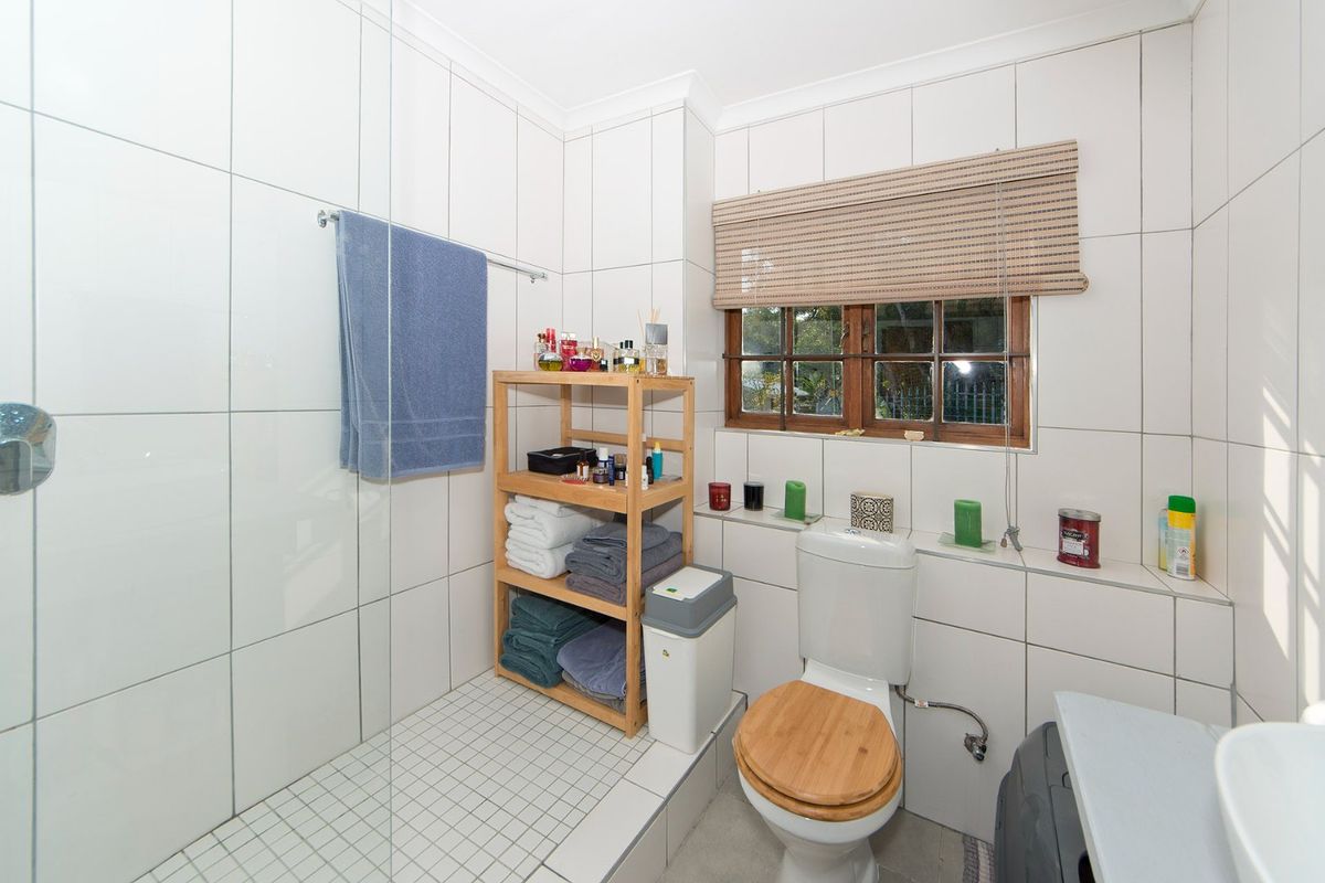 A functional bathroom featuring white wall tiles, a glass shower screen, a toilet, and a timber-framed window.