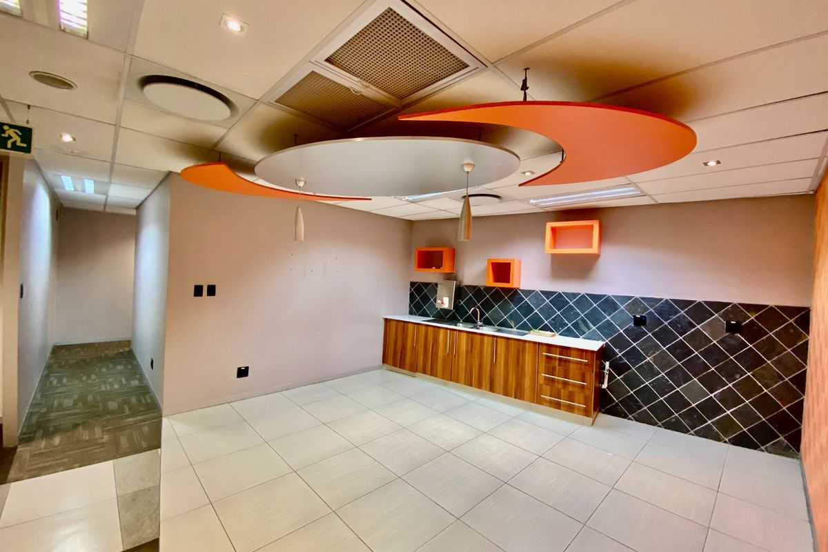 A clean, empty commercial kitchenette featuring wood-finish cabinetry, a dark tiled backsplash, and a decorative suspended ceiling feature with integrated lighting.