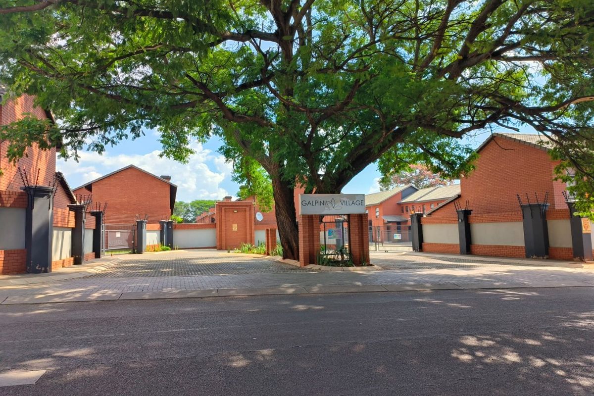 Street view of the entrance to Galpini Village, featuring a brick perimeter wall, security fencing, and a paved driveway under a large mature tree.