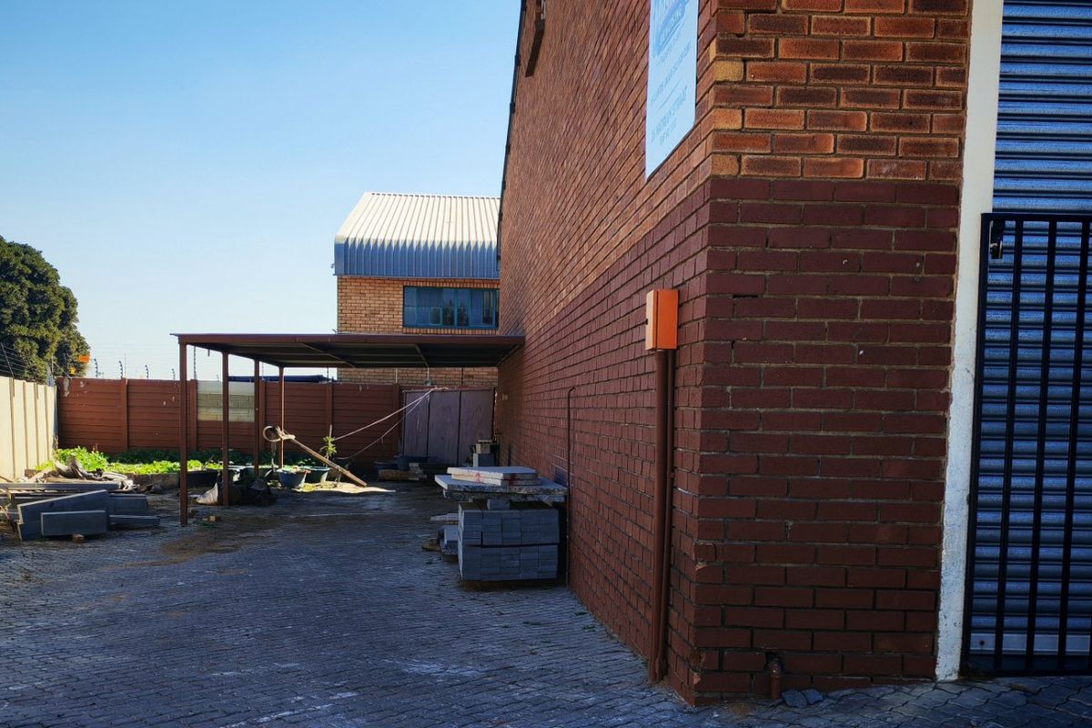 Rear exterior view of an industrial property featuring a brick facade, paved yard area, metal carport structure, and a secure roller shutter door.