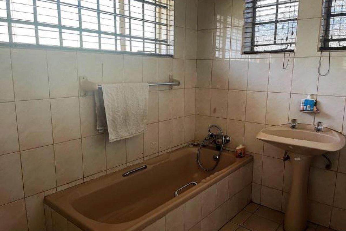 A dated bathroom featuring a built-in bathtub, pedestal basin, and full-height wall tiling with security bars on the windows.