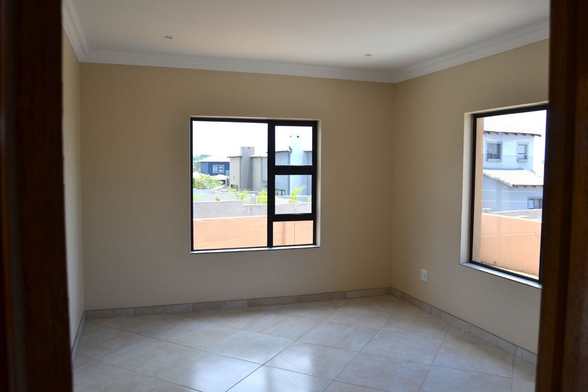 A bright, empty bedroom featuring neutral wall tones, tiled flooring, and modern aluminium-framed windows with views of the surrounding neighbourhood.