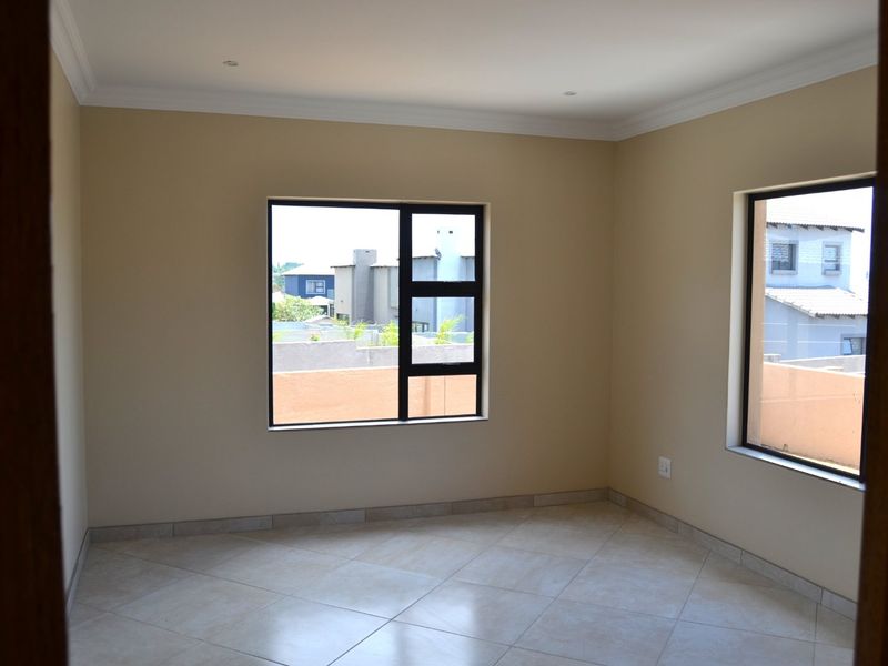 A bright, empty bedroom featuring neutral wall tones, tiled flooring, and modern aluminium-framed windows with views of the surrounding neighbourhood.
