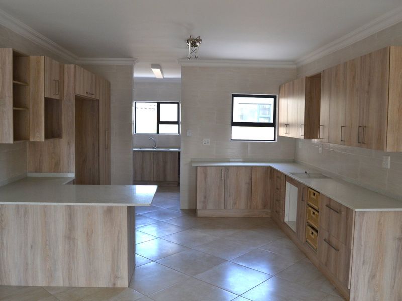 A spacious, modern kitchen featuring light wood-finish cabinetry, stone countertops, and tiled flooring, with an adjoining scullery area visible in the background.