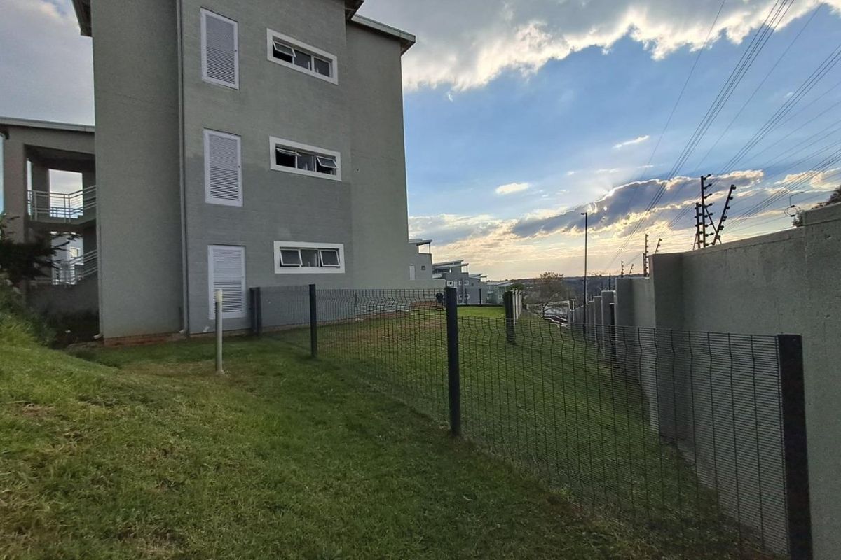 A view of the rear exterior of the apartment building, featuring a landscaped lawn, mesh perimeter fencing, and integrated security electric fencing.