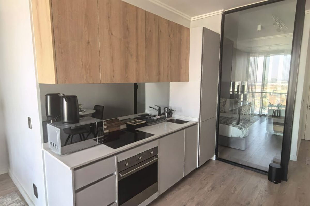 Modern compact kitchen featuring light wood-tone upper cabinetry, sleek grey lower cabinets, integrated oven, electric hob, and a contemporary glass partition wall.