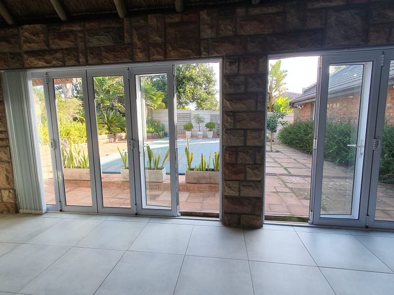 Spacious covered patio featuring stone-clad walls, large stacking glass doors, and tiled flooring, offering a seamless transition to the pool and garden area.