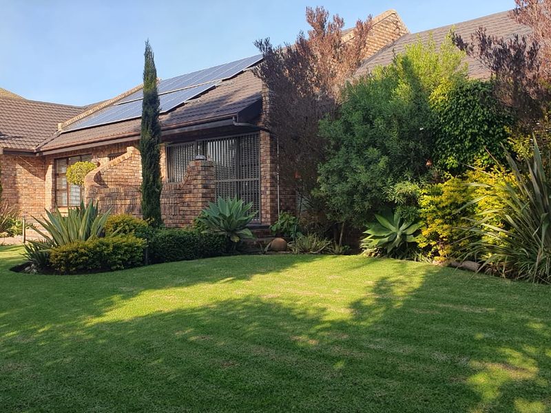 A well-maintained rear exterior view of a brick house featuring a lush green lawn, mature landscaping, and roof-mounted solar panels.