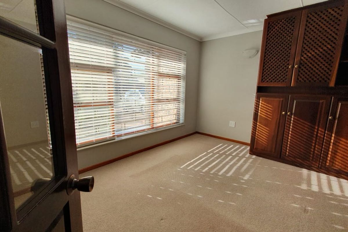 A bright, empty bedroom featuring neutral carpeting, large windows with venetian blinds, and substantial built-in wooden cabinetry.