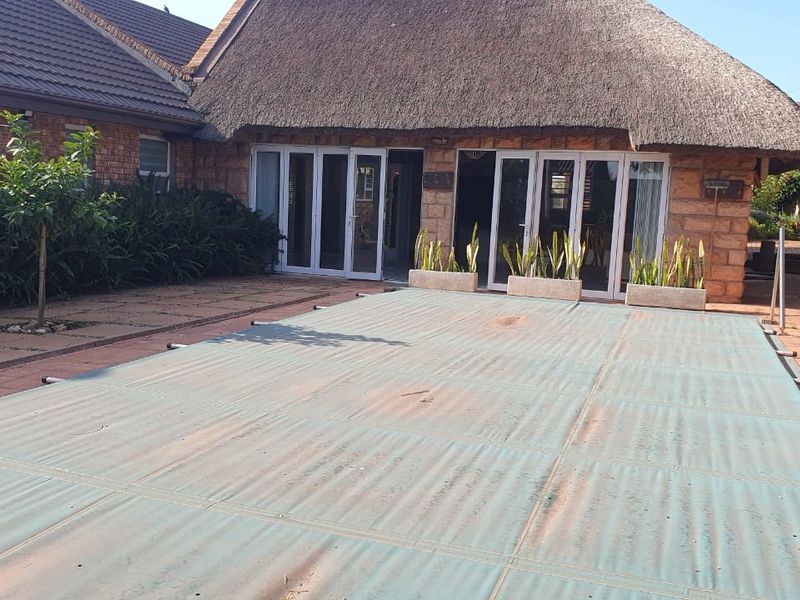 Outdoor pool area featuring a covered swimming pool, paved patio, and a traditional thatched-roof building with large sliding glass doors.