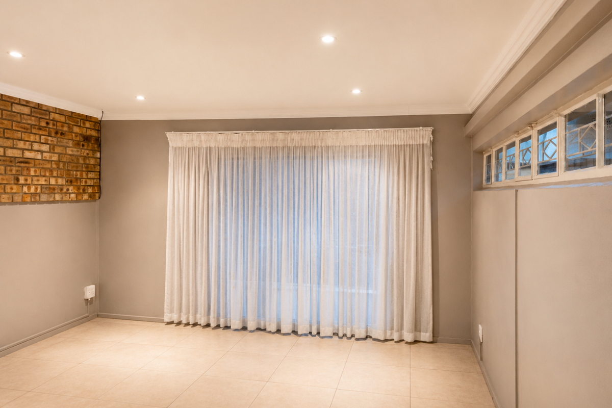 A bright, empty living room featuring tiled floors, an exposed brick accent wall, recessed lighting, and large windows with floor-to-ceiling curtains.