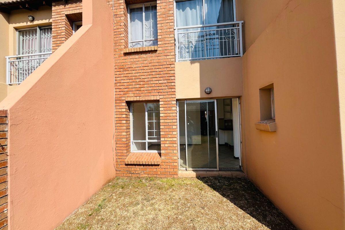 Rear view of an apartment unit featuring a small garden area, brick facade, sliding glass door access, and an upper-level balcony with security railings.