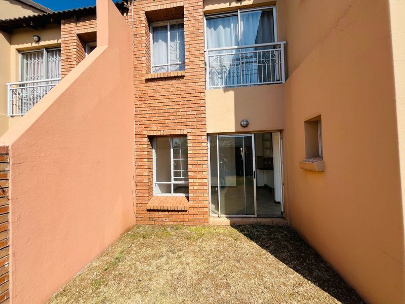 Rear view of an apartment unit featuring a small garden area, brick facade, sliding glass door access, and an upper-level balcony with security railings.