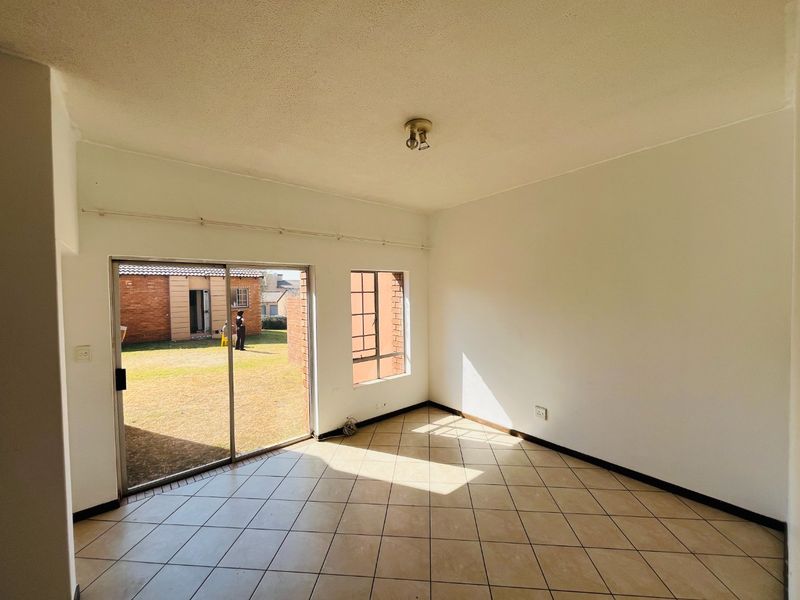 An empty, sunlit living room featuring tiled flooring, a sliding glass door leading to the garden, and large windows providing natural light.