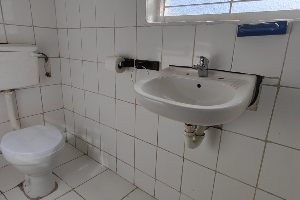 A dated bathroom featuring a wall-mounted basin, high-level cistern toilet, and fully tiled surfaces, requiring cosmetic updates and maintenance.