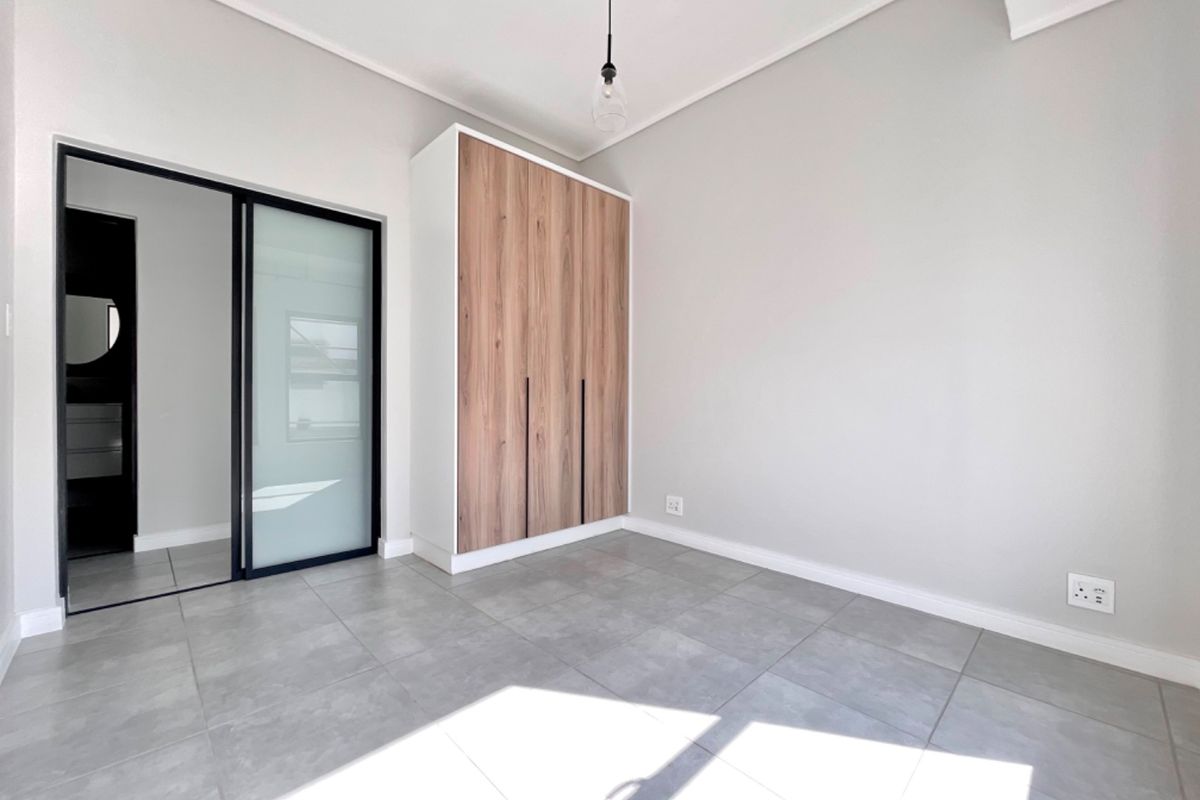 A bright, modern bedroom featuring neutral tiled flooring, a built-in wood-finish wardrobe, and a sleek sliding glass door leading to an adjacent space.