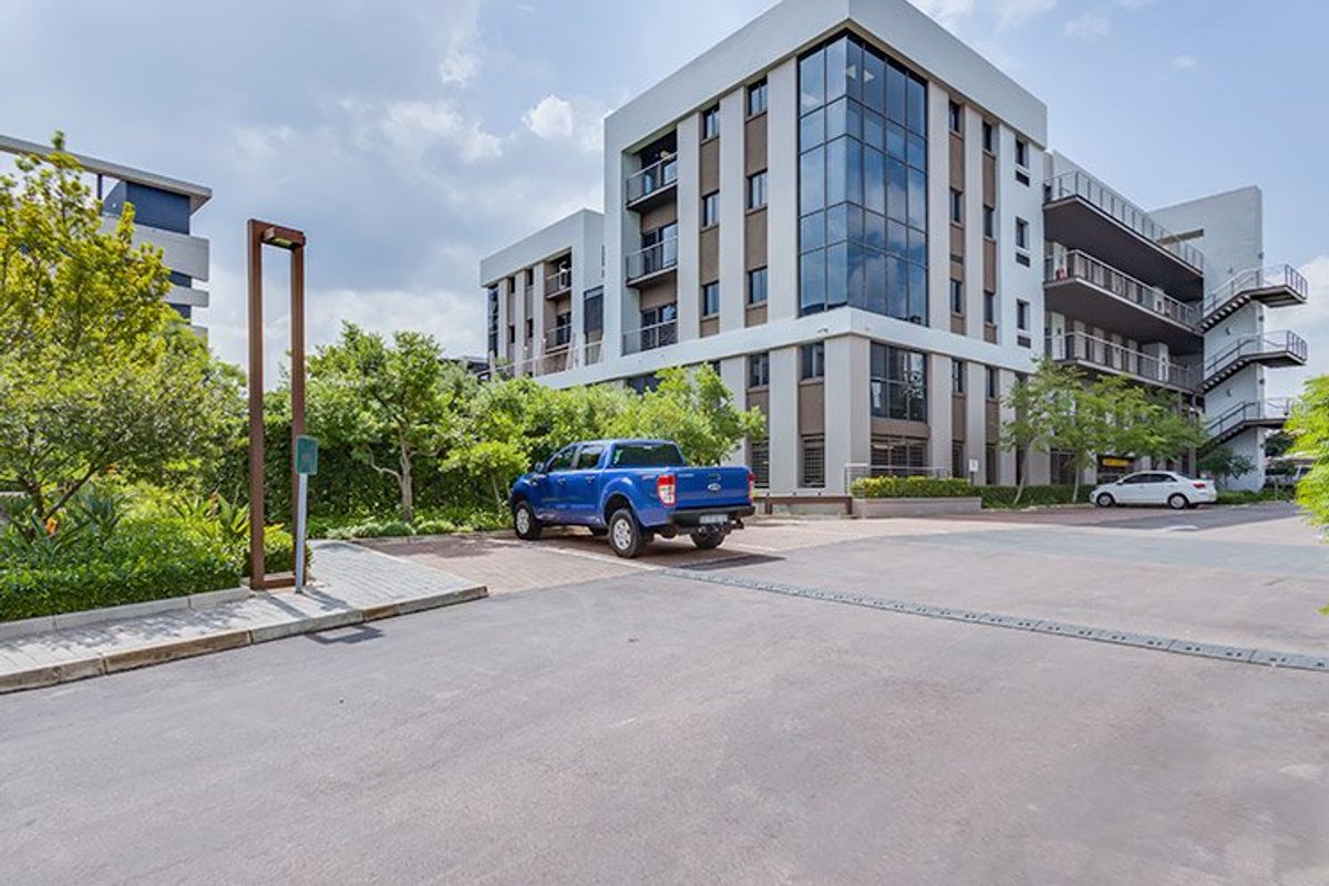 A modern multi-story commercial building featuring glass facade elements, balconies, and an adjacent paved parking area with landscaped surroundings.