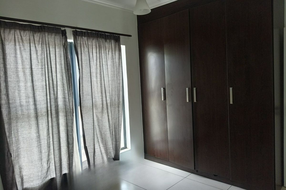 A clean, empty bedroom featuring dark wood built-in wardrobes, neutral tiled flooring, and large windows with curtains.