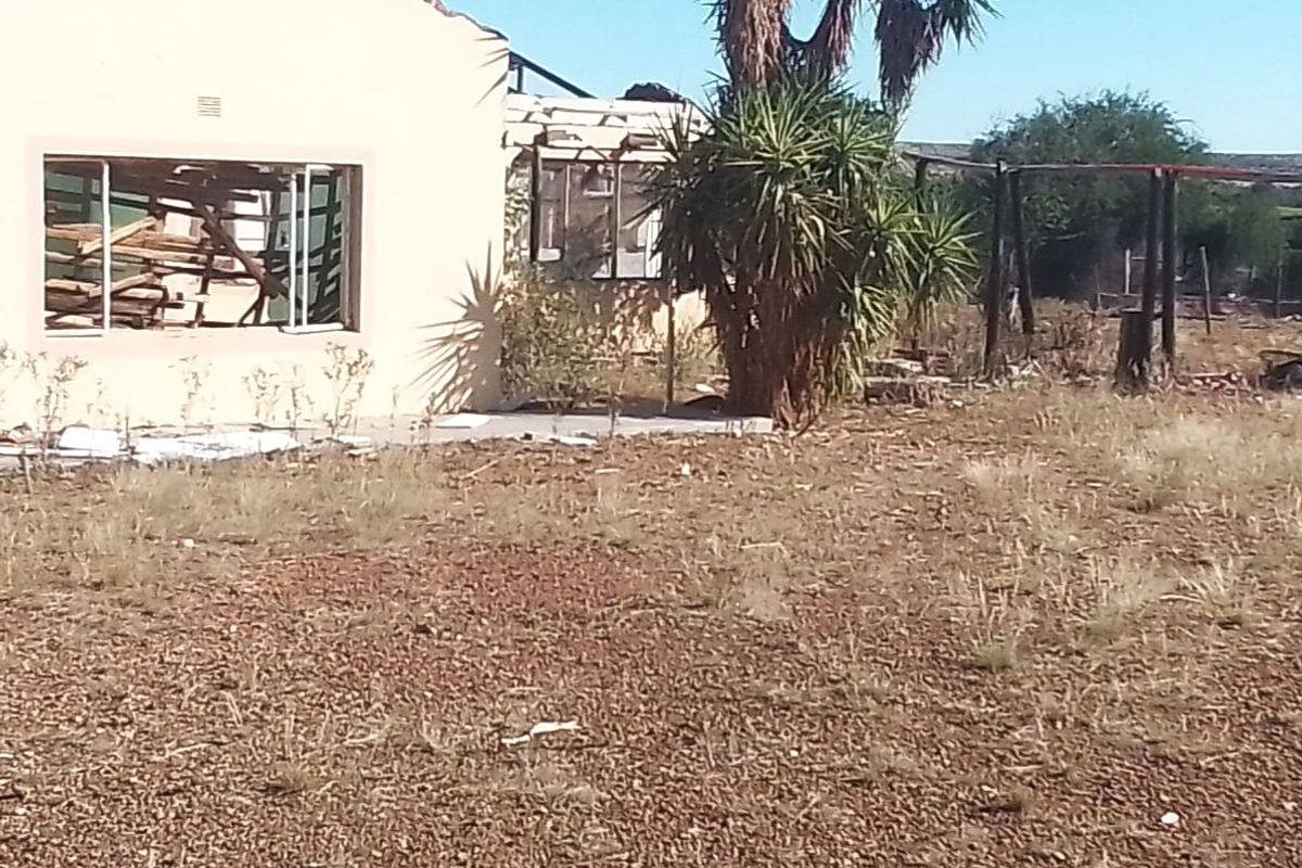 Rear view of a severely damaged residential structure showing collapsed roofing and broken windows on a large, unmaintained plot of land.