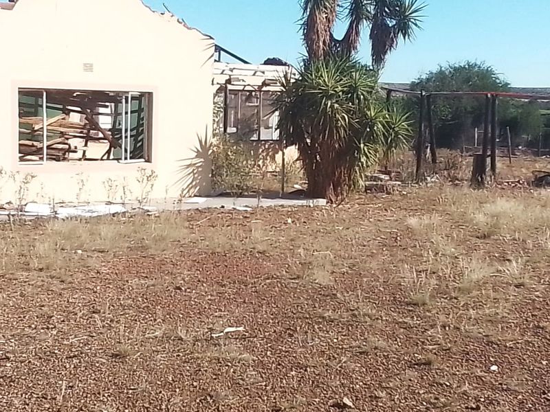 Rear view of a severely damaged residential structure showing collapsed roofing and broken windows on a large, unmaintained plot of land.