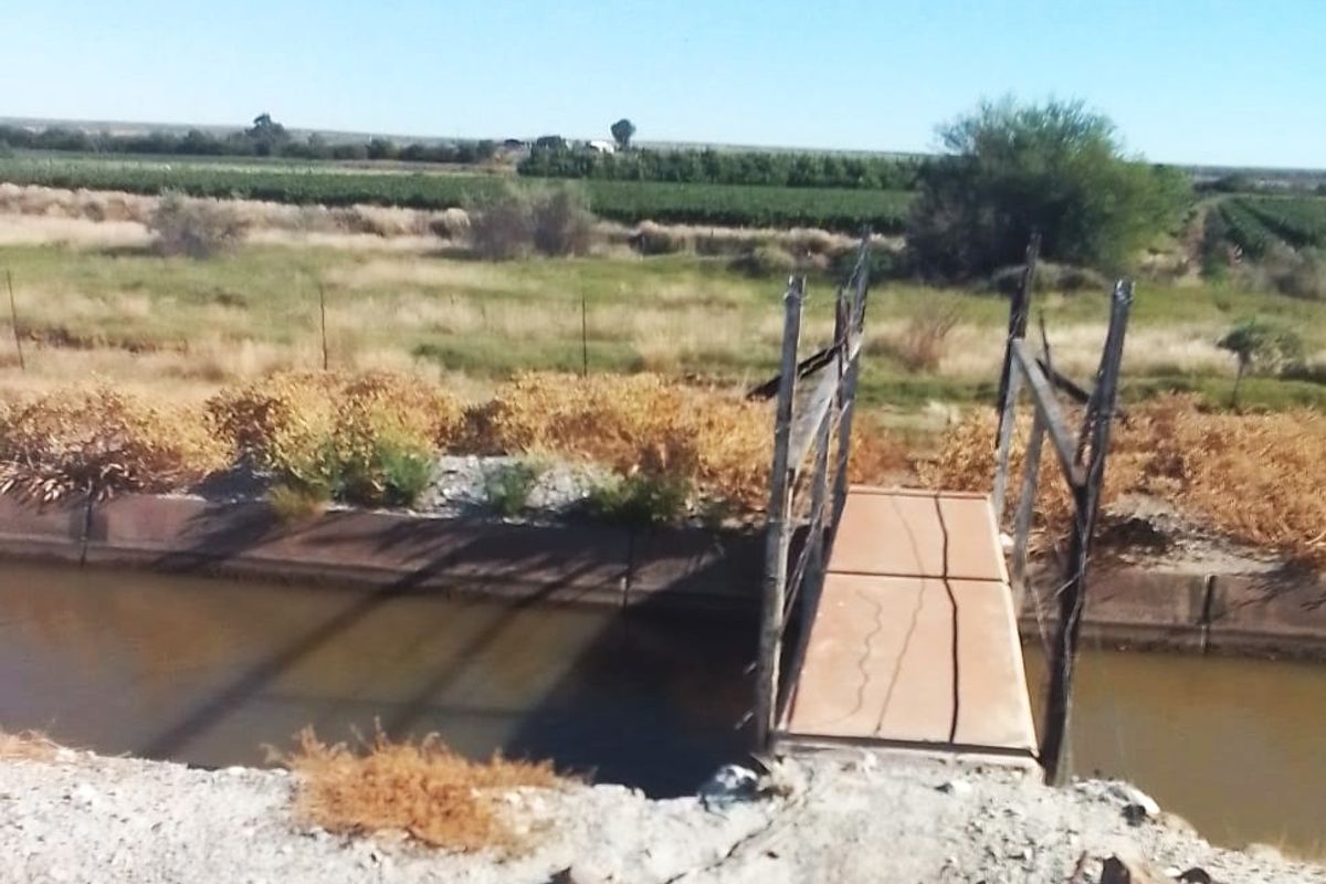 A view of a rural property featuring an irrigation canal crossed by a small, weathered pedestrian bridge with surrounding open land.
