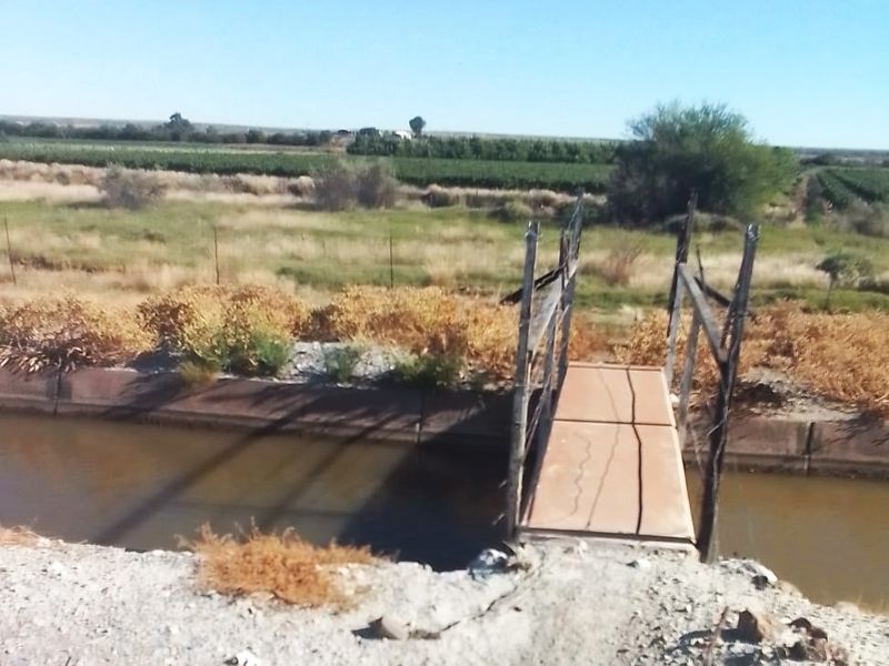 A view of a rural property featuring an irrigation canal crossed by a small, weathered pedestrian bridge with surrounding open land.