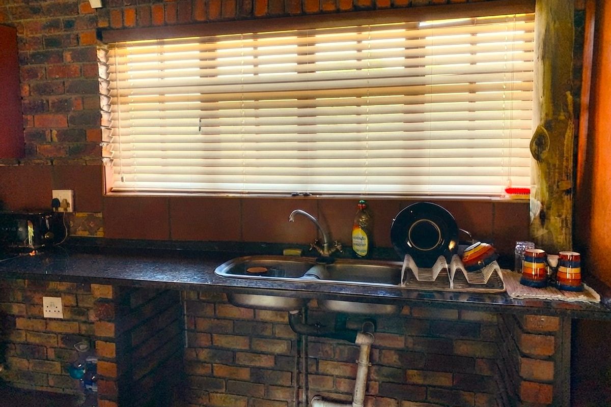 A rustic kitchen area featuring exposed brick walls, a granite countertop, and a double bowl stainless steel sink set beneath a window with blinds.