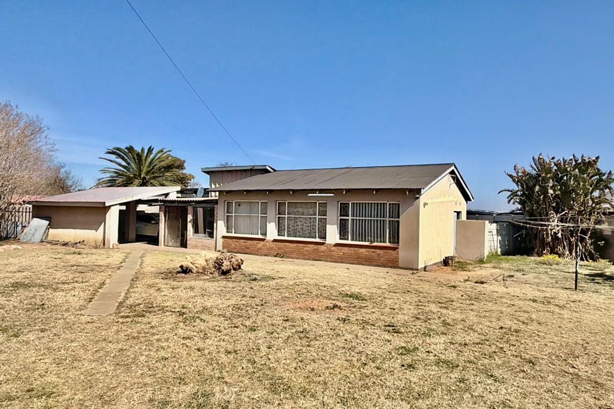 Rear view of a single-story house featuring a corrugated metal roof, brick facade, and a large backyard area with a concrete pathway.
