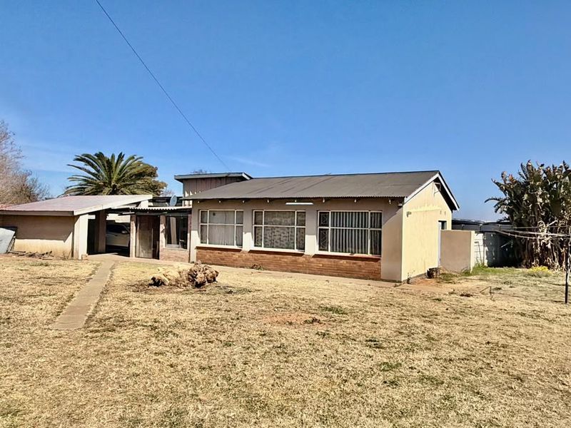 Rear view of a single-story house featuring a corrugated metal roof, brick facade, and a large backyard area with a concrete pathway.