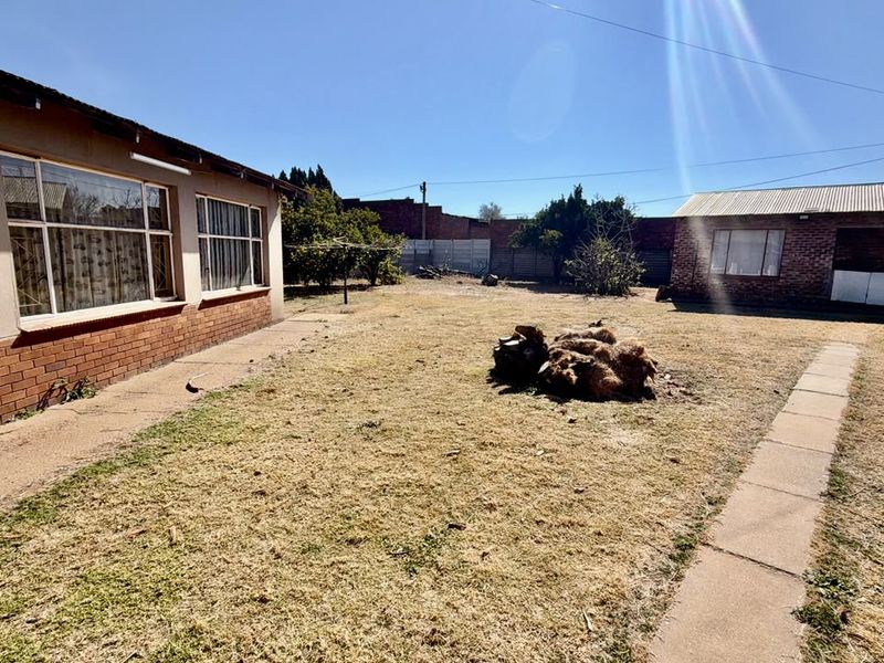 Spacious rear garden area featuring a brick-built main house, concrete pathways, and a detached outbuilding under a metal roof.