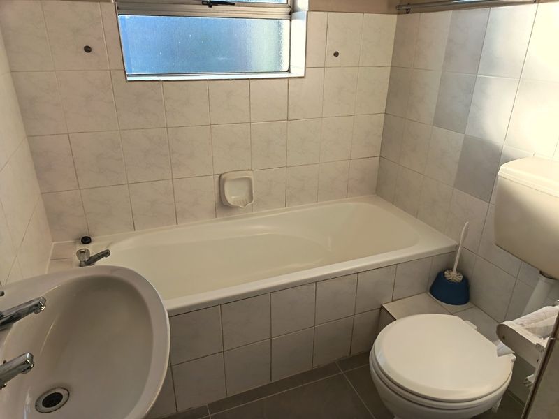 A functional bathroom featuring a standard bathtub, pedestal basin, and toilet with neutral wall and floor tiling.