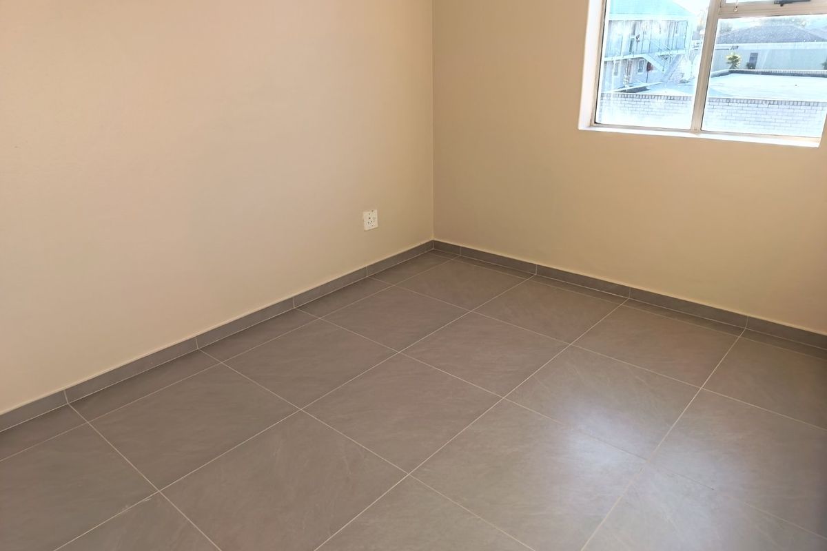 A clean, empty bedroom featuring neutral wall paint, modern grey floor tiles, and an aluminium-framed window providing natural light.