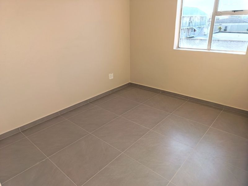 A clean, empty bedroom featuring neutral wall paint, modern grey floor tiles, and an aluminium-framed window providing natural light.