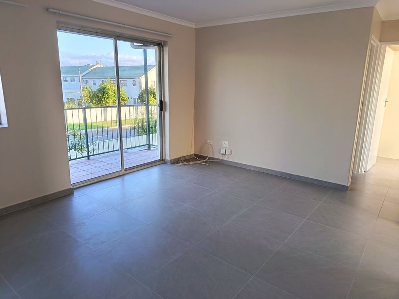 A bright, empty living room featuring modern grey floor tiles, neutral walls, and a sliding glass door leading to a private balcony.