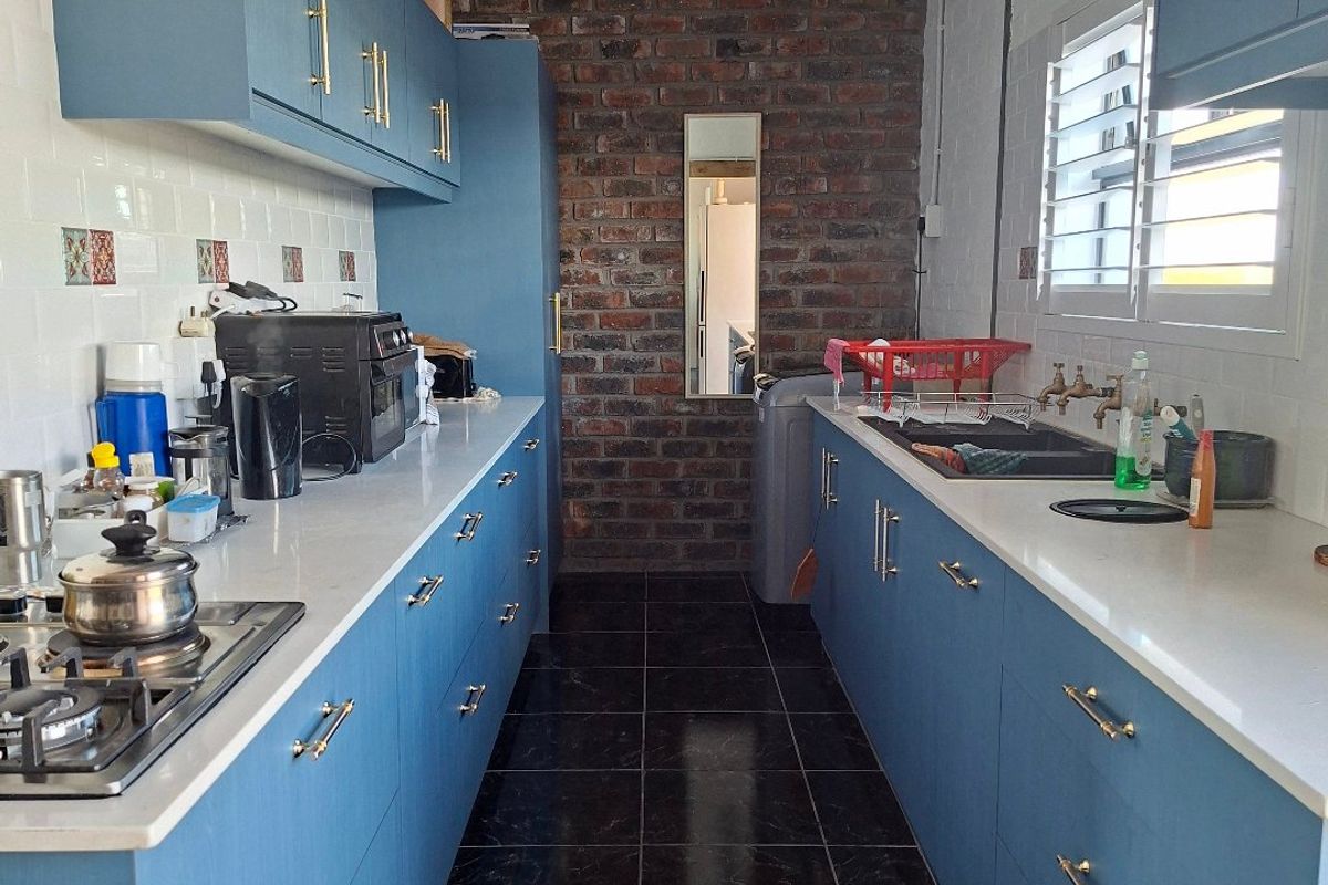 A modern galley-style kitchen featuring blue cabinetry, stone countertops, a gas hob, and a striking exposed brick feature wall with dark tiled flooring.