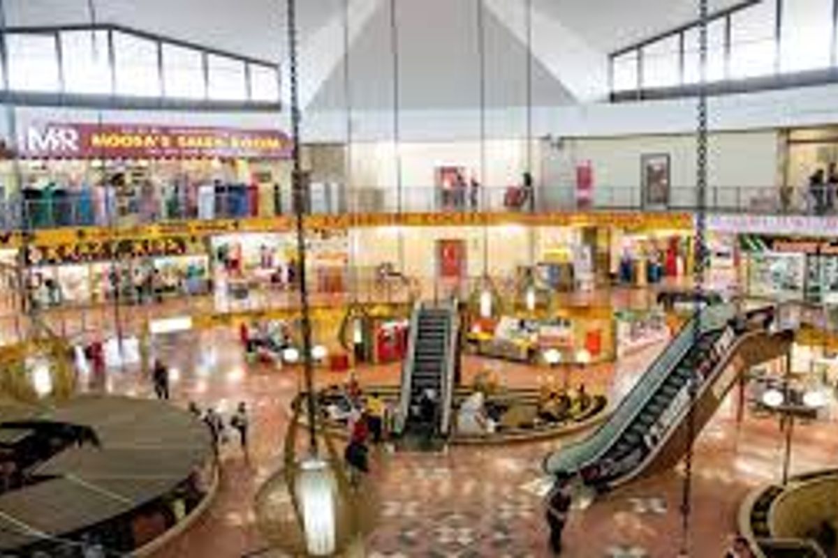 A multi-level commercial atrium featuring central escalators, mezzanine levels with retail storefronts, and large suspended pendant lighting fixtures.