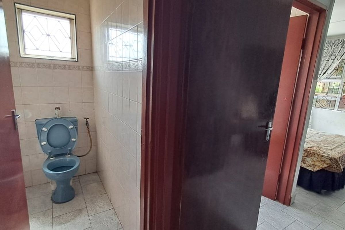 A small, dated bathroom featuring ceramic wall and floor tiles, a blue toilet, and security bars on the window.