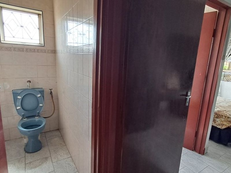 A small, dated bathroom featuring ceramic wall and floor tiles, a blue toilet, and security bars on the window.