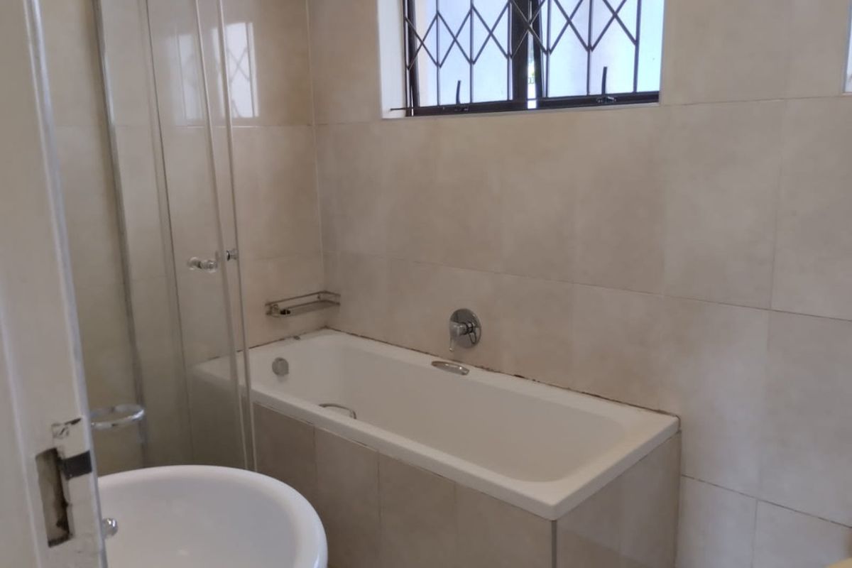 A functional bathroom featuring a built-in bathtub with a glass shower screen, neutral wall tiling, and a window with a security gate.