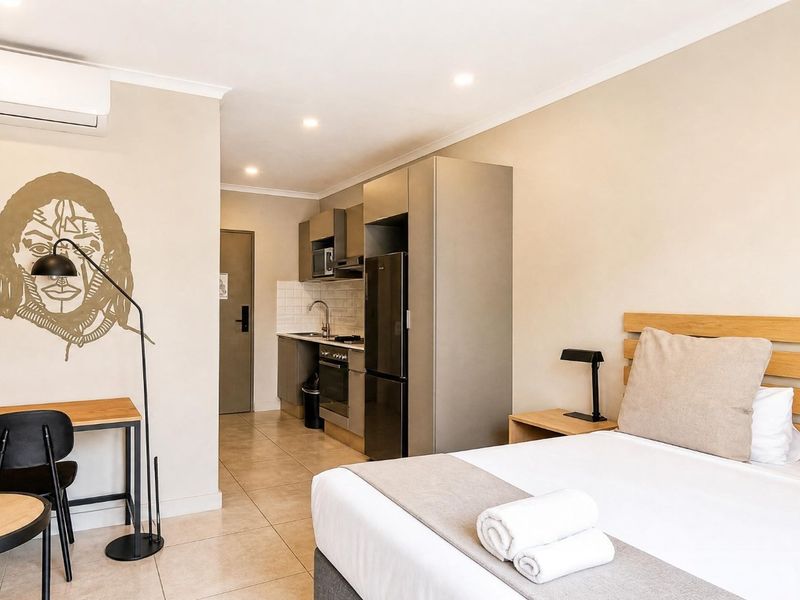 Modern open-plan studio apartment featuring a compact kitchenette with built-in appliances, tiled flooring, and a wall-mounted air conditioning unit.