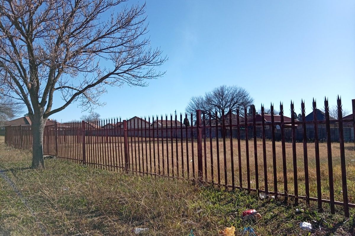A vacant plot of land enclosed by a metal spike perimeter fence, featuring dry grass and surrounding residential properties.