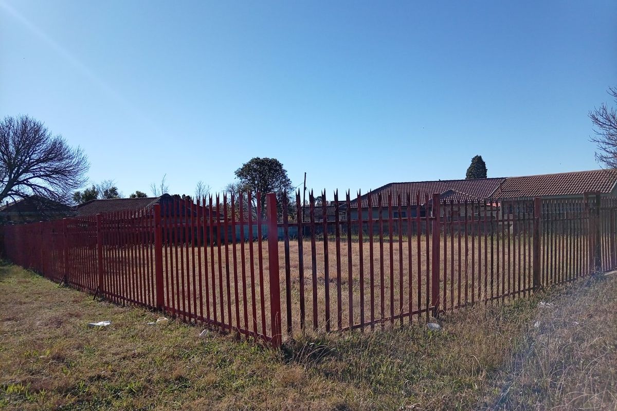 A vacant residential plot enclosed by metal spike perimeter fencing, situated in a suburban neighborhood setting.