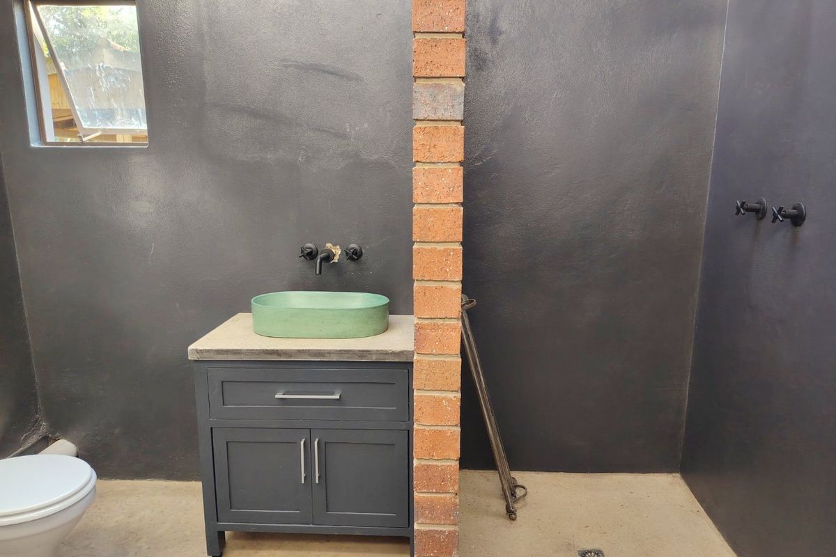 A modern industrial-style bathroom featuring a dark vanity with a concrete top, vessel sink, and an exposed brick pillar separating the toilet and shower area.