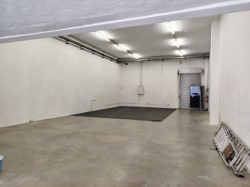 Spacious industrial utility area featuring concrete flooring, exposed electrical conduit, fluorescent lighting, and a secure roller shutter door access.