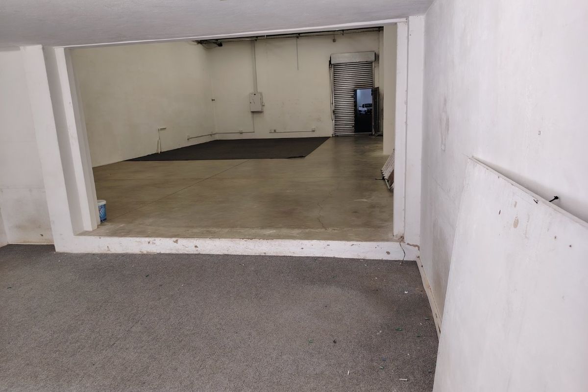 An open-plan industrial utility space featuring concrete flooring, a roller shutter door, and basic electrical infrastructure, suitable for storage or workshop use.
