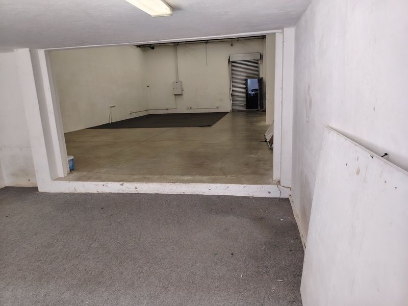 An open-plan industrial utility space featuring concrete flooring, a roller shutter door, and basic electrical infrastructure, suitable for storage or workshop use.